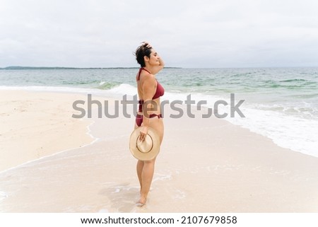 Full length view of latin american woman on tropical island seashore. Panoramic view of woman in bikini with hat on her hand on paradisiacal beach. People traveling and holidays concept