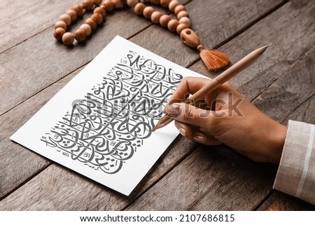 Arabic calligraphist writing on paper sheet against wooden background Royalty-Free Stock Photo #2107686815