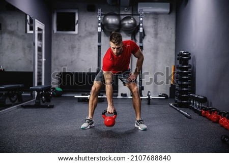 Cross fit training with kettle bell lifting. An attractive and muscular man in sportswear pumps his muscles and lifts a kettle bell in a modern gym. Strength, strong movement, arm and back workout Royalty-Free Stock Photo #2107688840