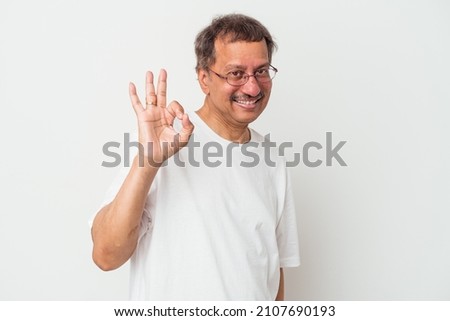 Middle aged indian man isolated on white background winks an eye and holds an okay gesture with hand.