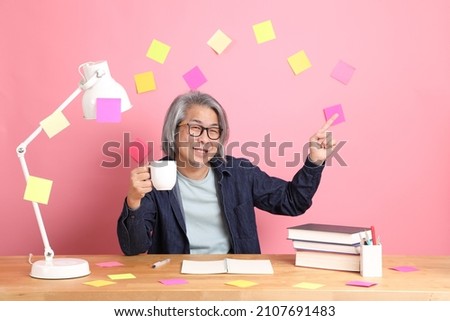 The senior Asian man sitting at the desk with many books with pink wall.