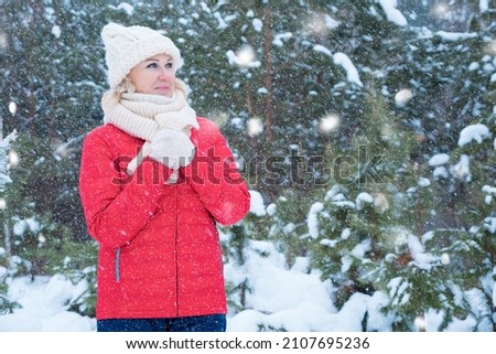 Blonde woman dressed in warm seasonal clothes stands against lush coniferous trees covered with snow posing for photo in winter forest with snowdrifts.