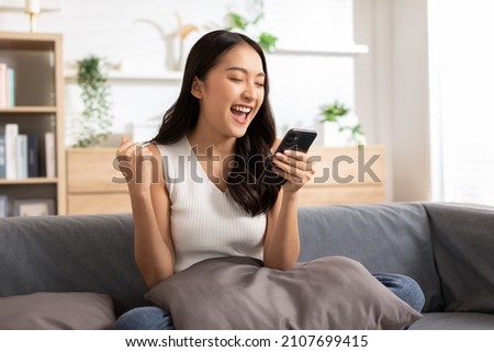 Millennial asian young woman looking mobile phone laughing with good news or discount voucher for shopping online at home.Happy and cheerful woman looking on cellphone app read message feel excited Royalty-Free Stock Photo #2107699415