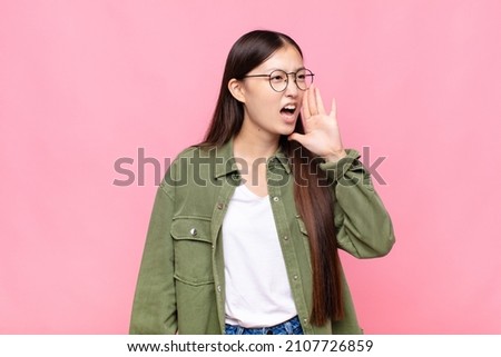 asian young woman yelling loudly and angrily to copy space on the side, with hand next to mouth