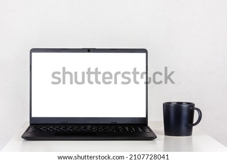 a white mockup on a laptop screen and a black mug of tea on a table against a white wall with a copy space