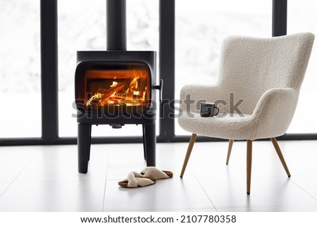 Cozy living space by the burning fireplace with chair, cup and slippers on background of snowy landscape. Idea of home comfort and winter mood Royalty-Free Stock Photo #2107780358
