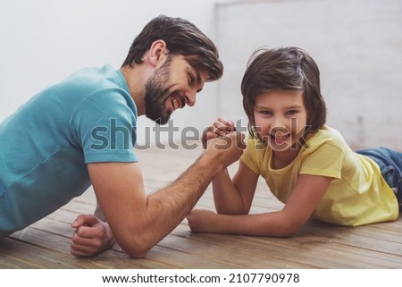 Handsome young father in casual clothes and his cute little son competing in arm wrestling while lying on a wooden floor in the room