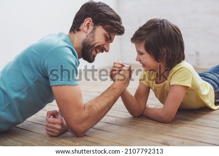 Handsome young father in casual clothes and his cute little son competing in arm wrestling while lying on a wooden floor in the room