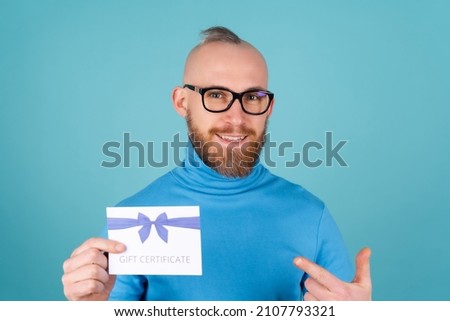 A young man with a red beard in a turtleneck and glasses on a blue background holds a gift certificate, smiles cheerfully, perky mood