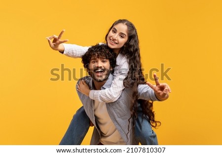Romantic indian couple having fun together, showing V-sign gesture, happy millennial guy piggybacking his girlfriend, joyful lady riding on man's back, yellow background
