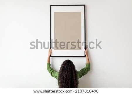Unrecognizable Female Hanging Blank Poster In Frame On White Wall Standing Back To Camera Indoors. Modern Art And Decor For Domestic Interior Concept. Mockup