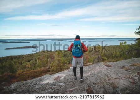 hiker wearing a jacket and carrying a backpack, standing on a rock watching Lake Jatkonjarvi at sunset in Koli National Park, eastern Finland. A man aged 24 wearing sports clothes. Active lifestyle. Royalty-Free Stock Photo #2107815962