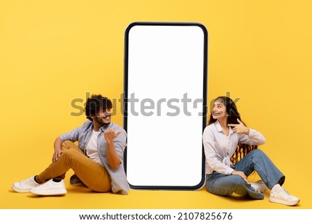 Happy indian couple sitting near big smartphone with blank white screen, demonstrating copy space for app or ad design, posing over yellow background, mockup banner Royalty-Free Stock Photo #2107825676