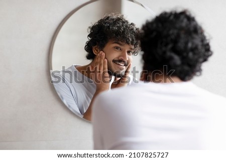 Attractive bearded indian man touching face applying moisturizer on face, standing near mirror in modern bathroom. Male facial skincare routine Royalty-Free Stock Photo #2107825727