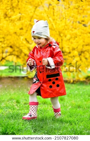 A cute little girl in a red jacket walks along the green grass against the background of a yellow bush.