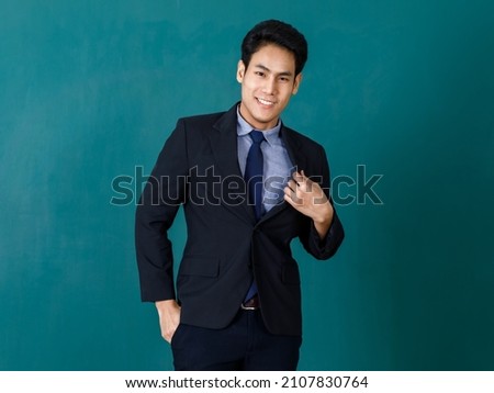 Portrait studio shot of Asian young professional successful male teacher in black formal suit and necktie standing smiling look at camera in front green blackboard background in classroom at school.