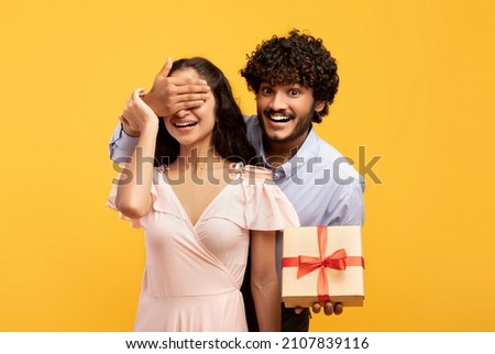 Happy anniversary. Loving indian man covering his girlfriend's eyes, greeting her with wrapped gift box, posing over yellow studio background, celebrating special day Royalty-Free Stock Photo #2107839116