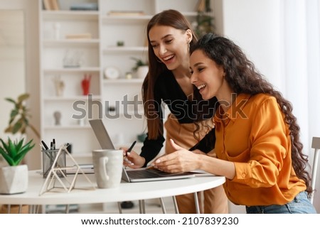 Two Happy Ladies Using Laptop Computer Working And Learning Online Together Or Making Video Call Sitting At Desk In Modern Office. E-Learning. Females Entrepreneurship Career Concept. Side View
