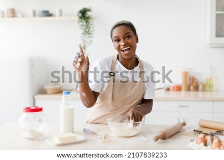 Laughing millennial african american woman in apron prepares dough for baking and raises finger up in light scandinavian kitchen interior. Got idea, cooking pizza, pie at home and facial expression Royalty-Free Stock Photo #2107839233