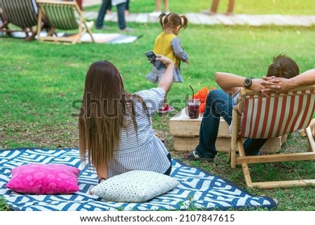 Back view of Asian woman sit to take selfie on mat near her lover on deck chair in garden. Summer vacation in green surroundings. Happy person outdoors relaxing in garden. Outdoor leisure.