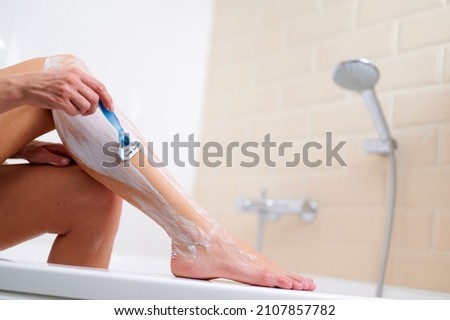 Caucasian woman shaving legs with razor blade in bathroom while sitting at the edge of the bathtub. Woman at bathroom. Royalty-Free Stock Photo #2107857782