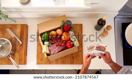 Overhead Of Man In Kitchen Holding Recipe Cards For Online Meal Food Recipe Kit Delivered To Home Royalty-Free Stock Photo #2107874567