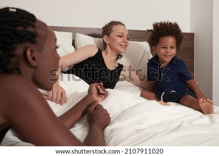 Boy with strabismus sitting in bed with his two mothers and watching movie or animated cartoon