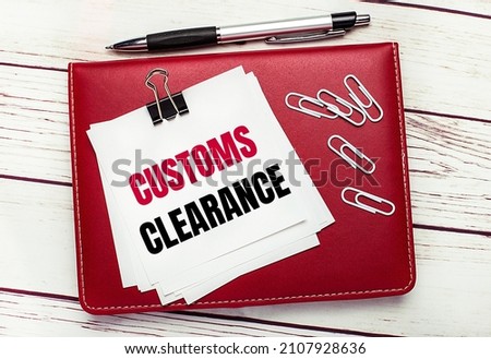 On a light wooden background, a burgundy pen and notebook. On the notebook has white paper clips and white paper with the text CUSTOMS CLEARANCE. Business concept