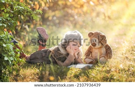 Little girl reading a book in a forest clearing with her teddy bear in autumn, fairy tale picture