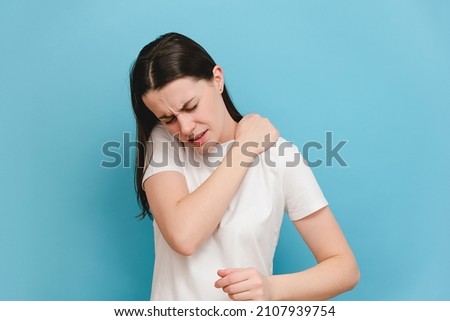 Tired upset young brunette woman massaging hurt stiff neck, fatigued sad brunette girl rubbing tensed muscles to relieve joint shoulder pain, isolated on blue studio background. Fibromyalgia concept Royalty-Free Stock Photo #2107939754