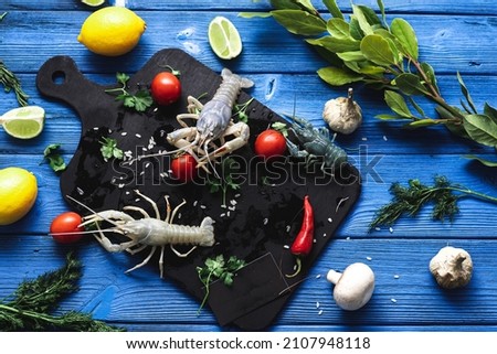 Live crayfish on a black cutting board, on a blue background of different colors next to vegetables and greens