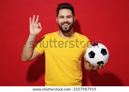 Charismatic stunning fun young bearded man football fan in yellow t-shirt cheer up support favorite team hold soccer ball showing okay ok gesture isolated on plain dark red background studio portrait