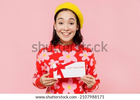 Smiling happy woman of Asian ethnicity 20s wear red knitted sweater beret hold gift certificate coupon voucher card for store isolated on plain pastel pink background. People lifestyle fashion concept