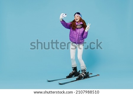 Full body skier woman in warm purple padded jacket ski goggles mask spend extreme weekend in mountains do selfie shot on mobile cell phone gesture show v-sign isolated on plain blue background studio.