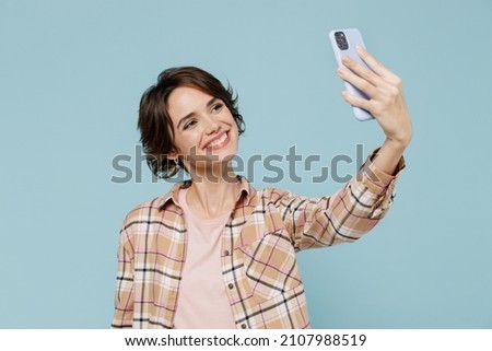 Young smiling woman 20s in brown shirt doing selfie shot on mobile cell phone post photo on social network isolated on pastel plain light blue color background studio portrait People lifestyle concept