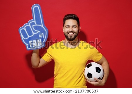 Blithesome young bearded man football fan in yellow t-shirt cheer up support favorite team look camera hold soccer ball fan foam glove finger up isolated on plain dark red background studio portrait Royalty-Free Stock Photo #2107988537