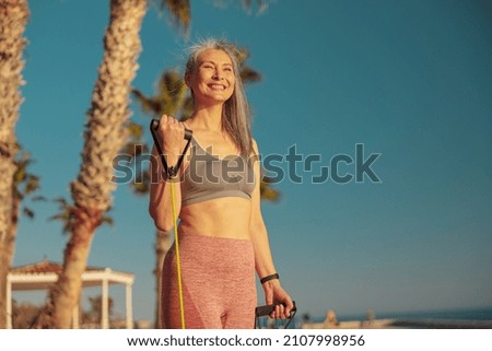 Happy Asian lady exercising using rubber bands bending her arm at elbow, standing against the background of palm trees, smiling