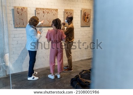 Young intercultural couple in masks listening to mature artist during presentation of his new artwork collection in gallery