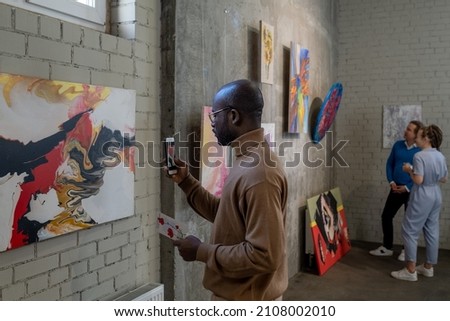 Young African male visitor of art gallery taking photo of painting on wall while enjoying exhibition of modern artists