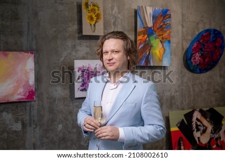 Contemporary mature man in elegant suit holding flute of champagne while standing against wall with collection of artworks