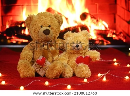 pair of teddy bears are sitting hugging against the background of a red knitted plaid and a garland of light bulbs and holding two red hearts in their paws
