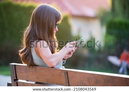 Young woman sitting on park bench browsing her cellphone outdoors on warm summer evening. Communication and mobile connection concept.