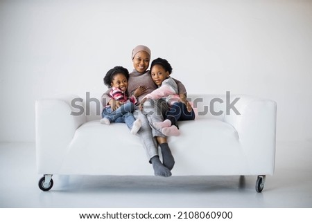 Beautiful african american muslim woman sitting on couch with her two pretty daughters and smiling on camera. Sisters embracing mother in studio. Royalty-Free Stock Photo #2108060900
