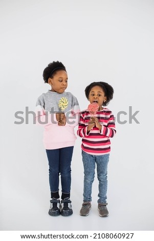 Cute african american sisters dressed in casual wear licking sweet colorful lollipops while standing over white studio background. Happy children enjoying sugar candies indoors.