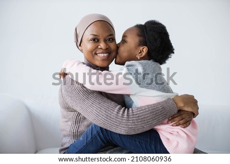 Charming african woman in headscarf smiling sincerely on camera while her pretty daughter gently kissing her. Happy mother with child relaxing on white couch in studio.