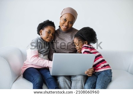 Caring african american muslim mother sitting with her two cute daughter on white couch and using portable laptop. Family time with modern technology.