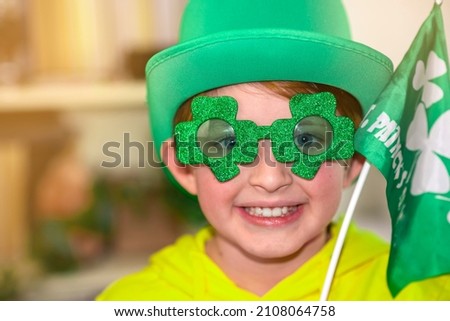 happy schoolboy on st. patrick day. wearing a green hat, wearing green clover-shaped glasses, waving a small green flag. at home