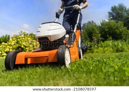 A young man is mowing a lawn with a lawn mower in his beautiful green floral summer garden. A professional gardener with a lawnmower cares for the grass in the backyard. Royalty-Free Stock Photo #2108078597