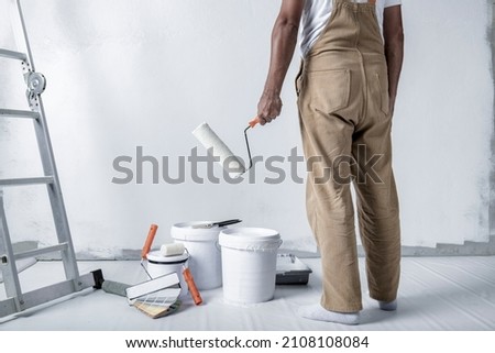 a man paints a white wall with a roller. Repair of the interior. Young male decorator painting a wall in the empty room, concept builder or painter in helmet with paint roller over the empty room. Royalty-Free Stock Photo #2108108084