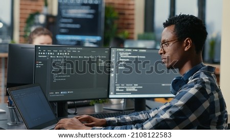 Sofware developer thinking while touching beard while typing on laptop sitting at desk with multiple screens parsing code. Focused database admin working with team coding in the background. Royalty-Free Stock Photo #2108122583